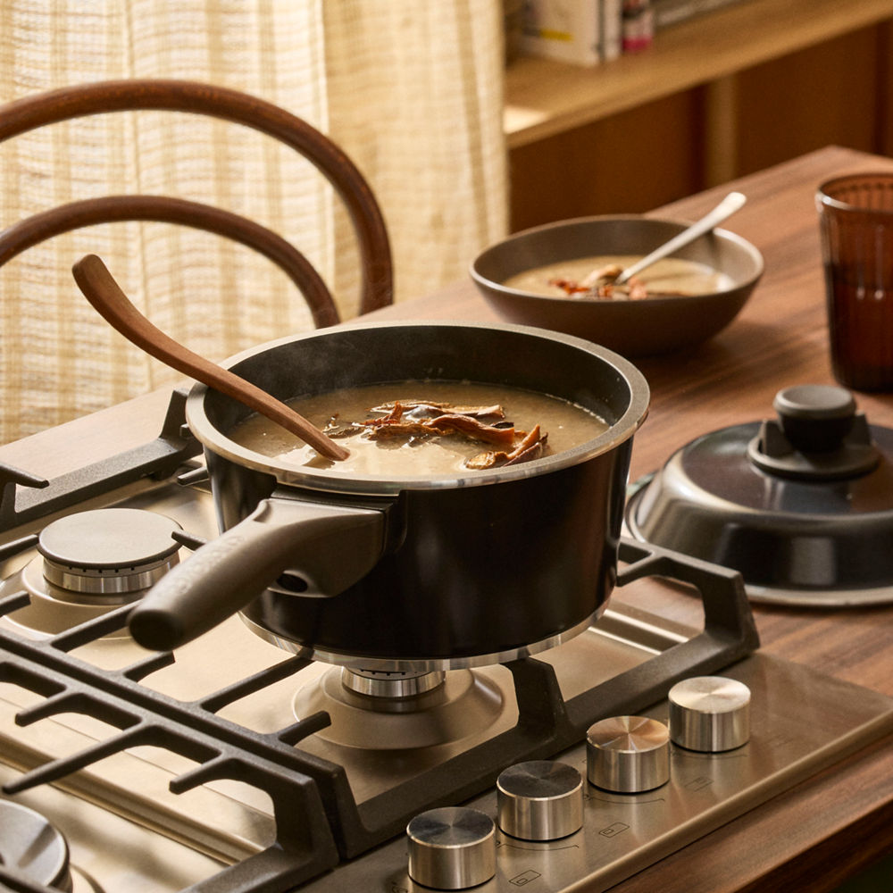 Black pot on a gas stove with a wooden spoon, surrounded by bowls and a cup on a wooden table.