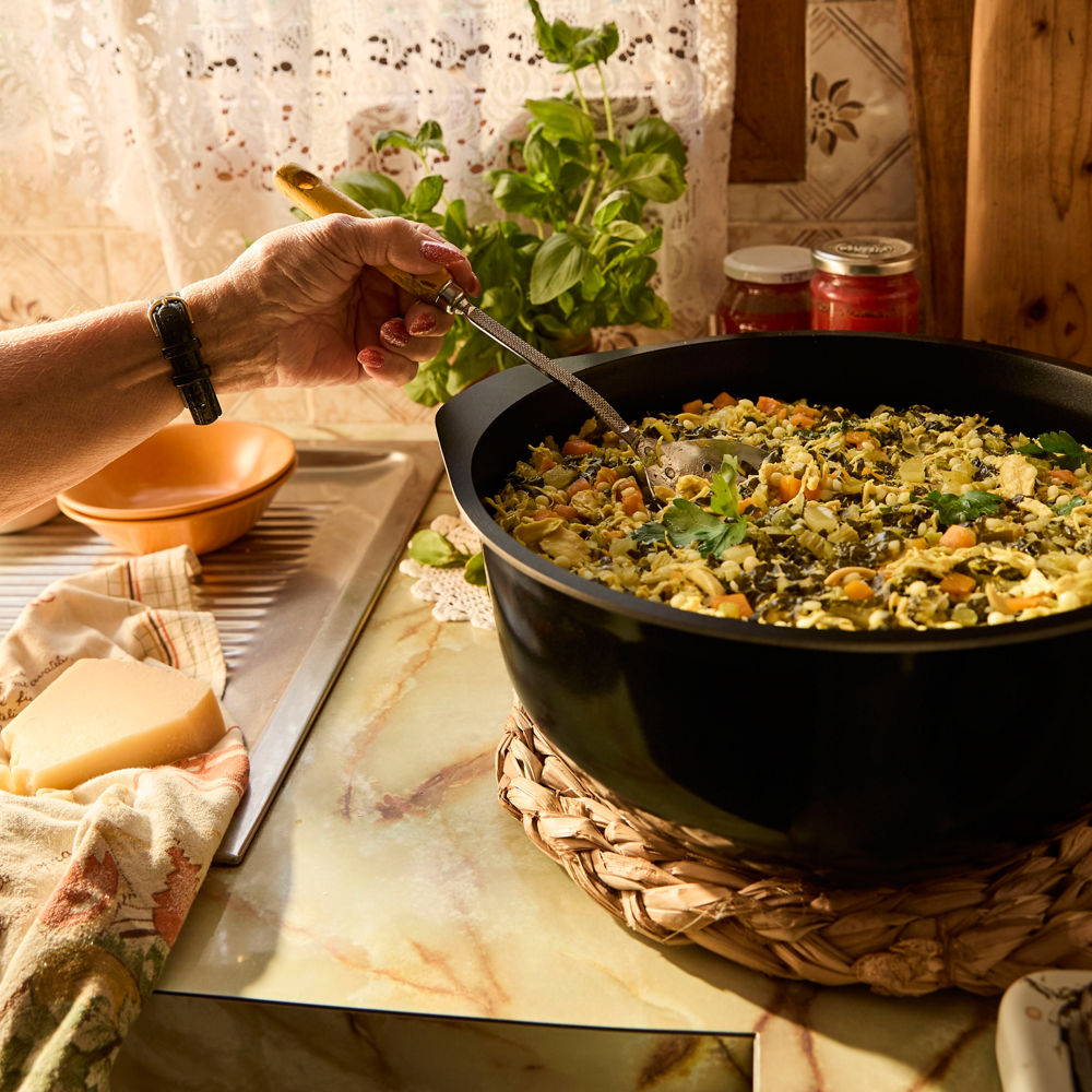 Person seasoning a dish in a black pan on a kitchen counter with herbs and cheese nearby.