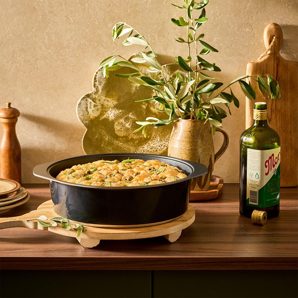 Baked dish in a black pan on a wooden surface with a plant and bottle in the background