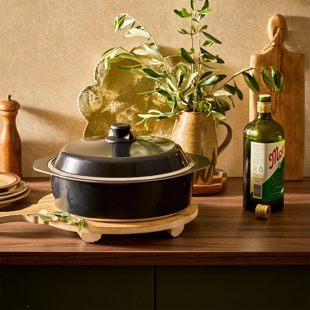 Black pot with a lid on a wooden stand in a kitchen setting with a bottle of olive oil and a plant.