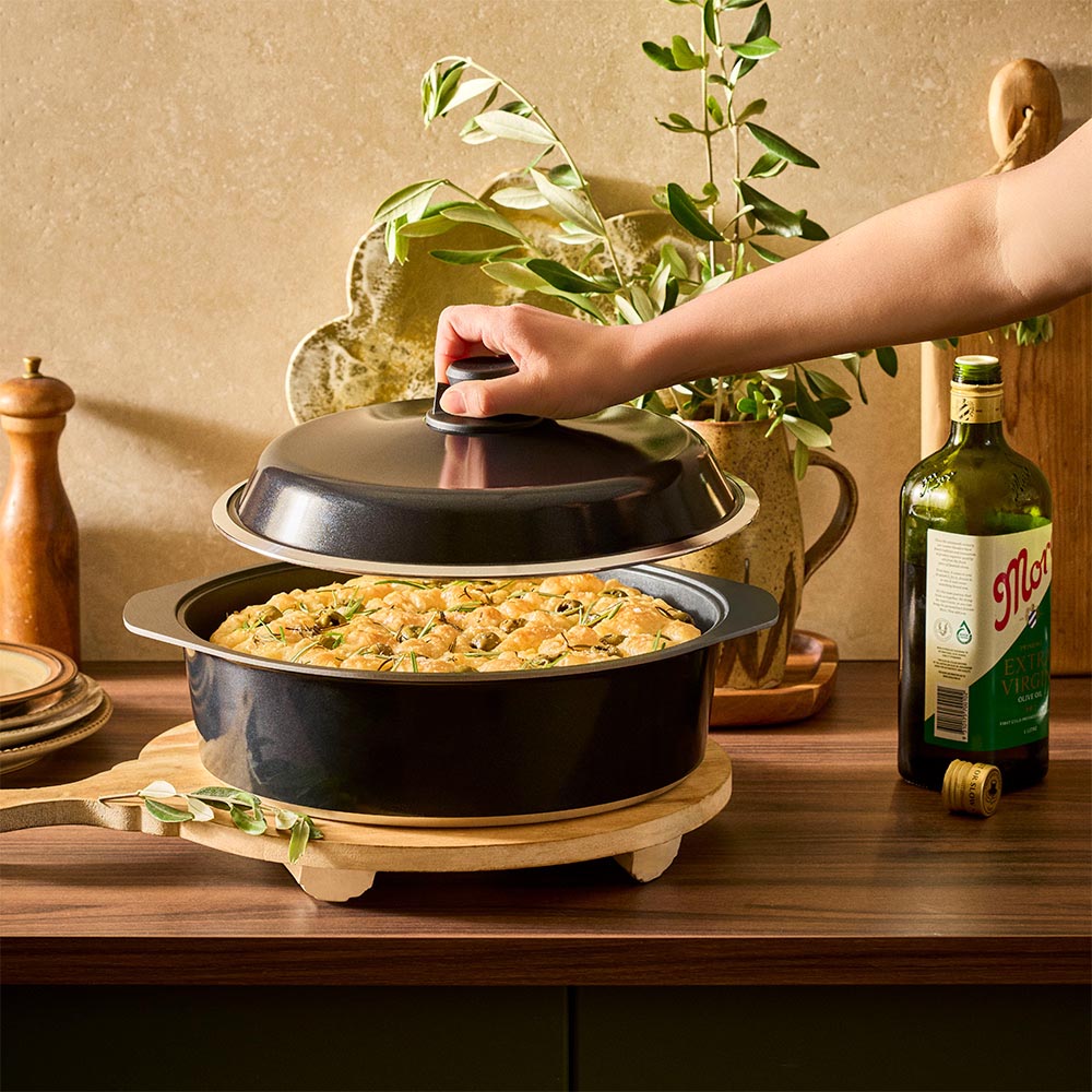 Person opening a black pot on a wooden surface with a bottle of olive oil and plant in the background.