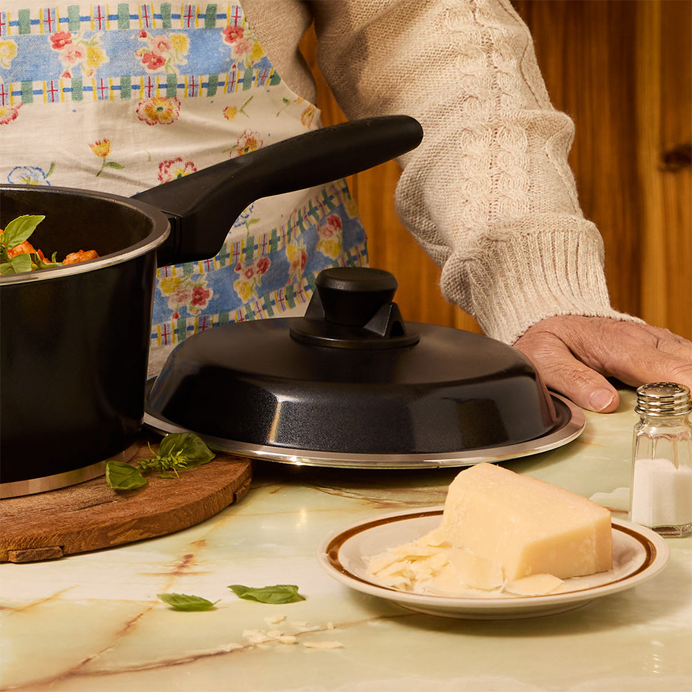 Person cooking with a pot and pan on a stove, next to a plate of cheese and a salt shaker.