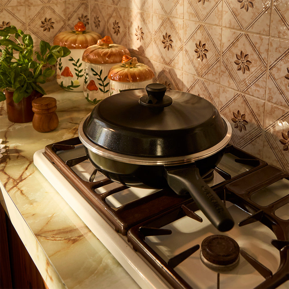 Black frying pan on a gas stove with decorative jars and plants in the background.