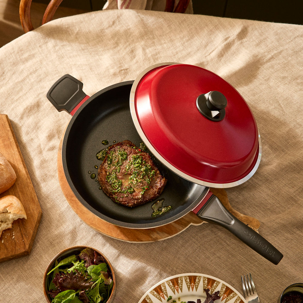 Red lid on a frying pan with a cooked steak on a table with bread and salad.