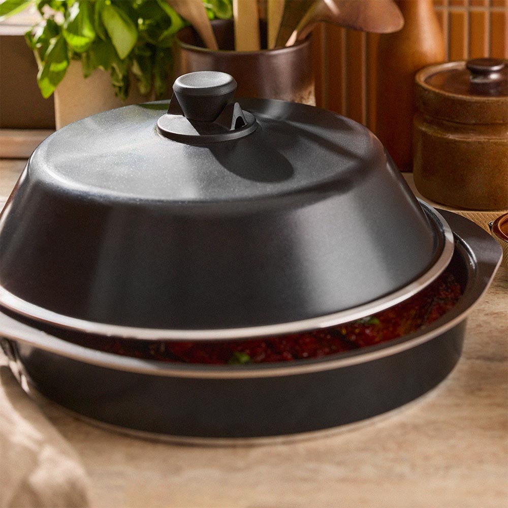 Black ceramic dish with lid on a wooden surface, surrounded by potted plants.