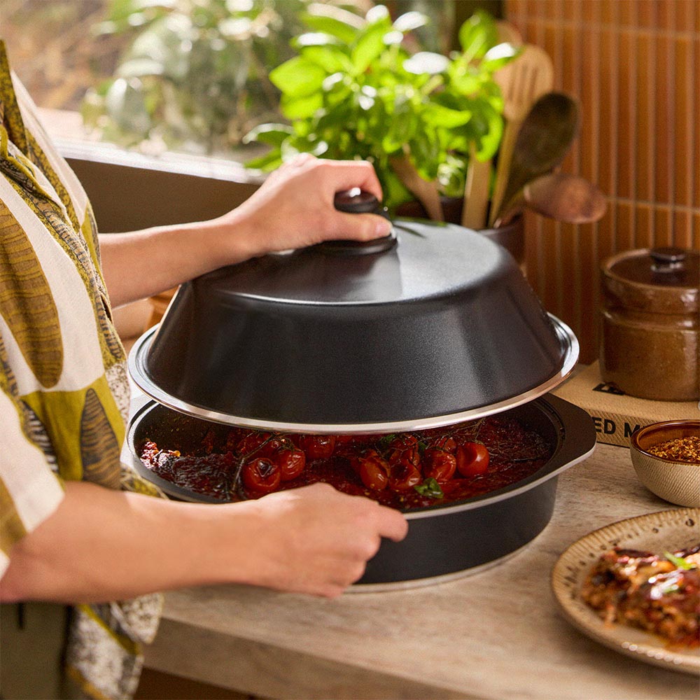 Person holding a black lid over a dish of food on a wooden table with plants in the background.