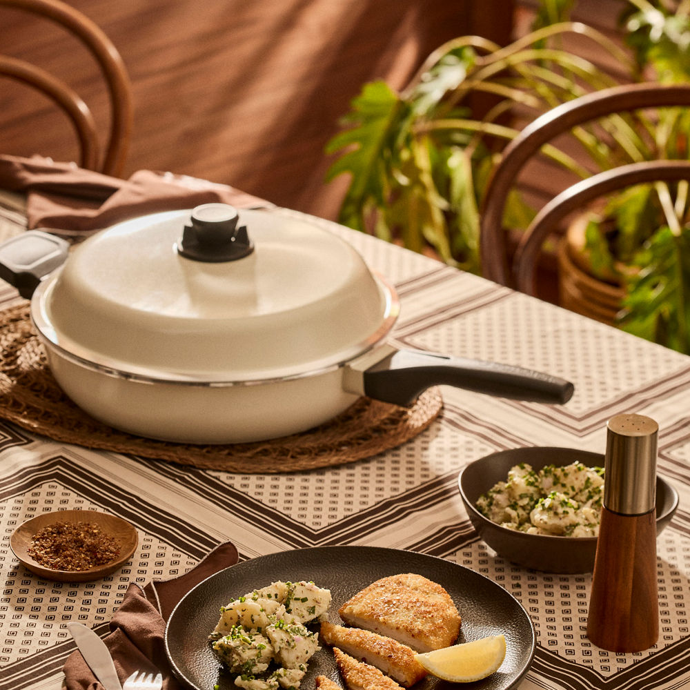 White pot with a black handle on a patterned tablecloth with food and a plant in the background